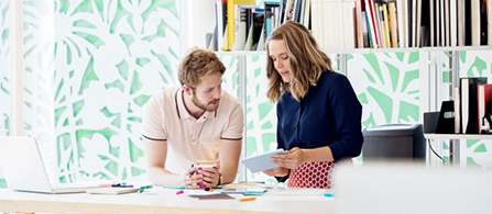 Two people work together at a white desk, looking at a tablet one of them holds. A green leaf-patterned wall and shelves of binders are behind them.