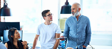 Three people in an office. One in a light blue shirt with a gray beard stands and speaks while gesturing; two colleagues sit nearby listening.