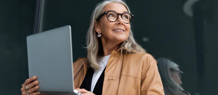 A person with gray hair wearing glasses and a tan jacket holds a laptop, looking upward with a slight smile. A dark reflective glass surface forms the background.
