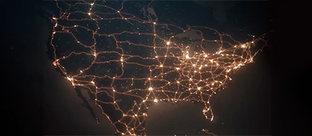 A nighttime aerial view of the United States with glowing orange network lines and clusters of city lights tracing routes across the country against a dark background.