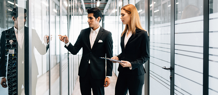 Two people stand in a server room with machines behind glass doors in a large, bright warehouse.