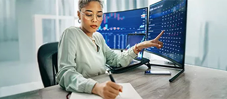 A business professional seated at their desk is writing notes in a notebook while looking away from their dual monitors. They have one finger pointed at the computer screen as they write. 