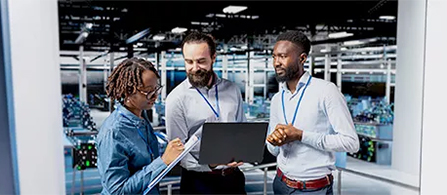 Three business professionals stand in a row, all looking at or gesturing toward a laptop the person in the middle holds between them. 