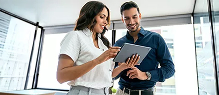 Two business professionals stand before a large opened window in a tall office building, with one holding a tablet for the other to look at.