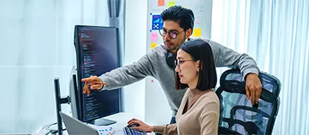Two business professionals are in discussion at work, one is standing and points to the computer monitor of the other who is sitting and typing.