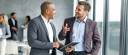 A small business gathering is taking place in a conference room with floor-to-ceiling windows taking up a whole wall. The focus are two business professionals smiling in conversation as one holds a tablet in their hand.