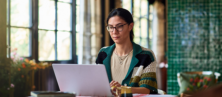 A woman sits at a table using a laptop computer in a bright indoor space. Large windows let in natural light, illuminating plants and decorative elements around her. She wears glasses and a patterned sweater, appearing concentrated on her work. The setting feels calm and cozy, suggesting remote work or study in a comfortable environment.
