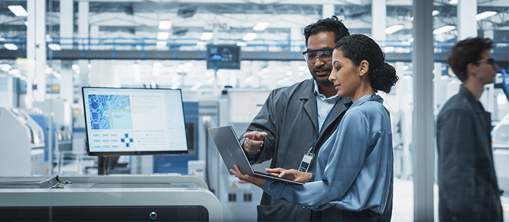 Two engineers stand in a high-tech manufacturing facility, reviewing information on a laptop computer. A large monitor beside them displays technical graphics and data, suggesting analysis or process monitoring. The environment is bright and industrial, with machinery and equipment visible in the background. The scene conveys teamwork, innovation, and advanced technology in an industrial setting.