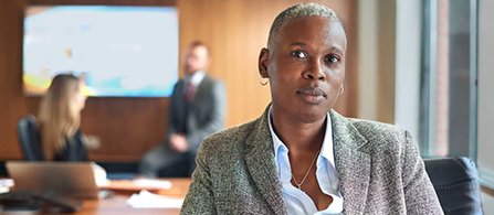 A person with short hair and small hoop earrings, wearing a gray tweed blazer over a white collared shirt, sits in a conference room and looks toward the camera with a calm, neutral expression. Two other people are blurred in the background near a wall-mounted screen and windows.