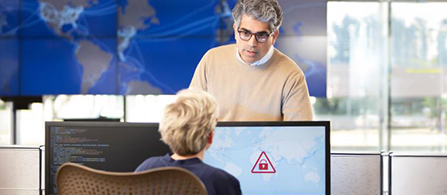 Two people work in an office facing each other across desks. One person with gray hair and glasses, wearing a tan sweater, stands and looks toward a monitor. In the foreground another person with short light hair sits facing a monitor that shows code and a red padlock warning icon. A blurred world map graphic appears on a screen in the background.