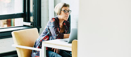 A person with short light hair, wearing glasses and a patterned red and blue jacket, sits at a white table beside a large window and works on a laptop, looking at the screen with a focused expression. A yellow upholstered armchair sits nearby and bright natural light fills the room.