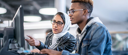 Two people sit side by side at a computer in a lab or office. One person, wearing a white head covering, glasses, and a dark jacket, points toward the screen with a focused expression. The other person, with short dark hair and glasses and wearing a denim jacket, looks at the screen. Computer equipment is blurred in the background.