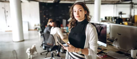 A person with long wavy hair, wearing a black sleeveless top and a white cardigan, stands in an open-plan office holding a smartphone and looks toward the camera with a relaxed expression. Behind them, other people work at desks with monitors in a loft-style space with columns and large windows.
