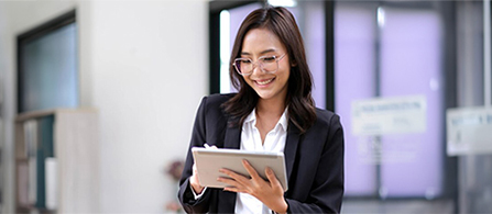A person in a dark blazer and glasses stands in a bright office holding a tablet, smiling while looking down at the screen. A purple-toned wall and glass partition are visible in the background.