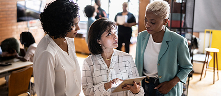 Three people stand together in a bright space looking at a tablet held by the person in the center, who speaks while the other two listen. Additional people are visible in the background at what appears to be a networking event or open office.