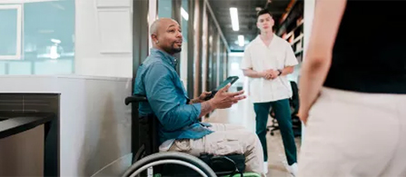 A person in a denim shirt sits and gestures with their hands while speaking, holding a smartphone, in a corridor of a modern facility. Another person in a white shirt stands further down the hallway, and a third is partially visible in the foreground.