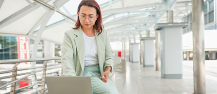 A person in a sage-green blazer and glasses sits on a bench at an outdoor transit platform or walkway, working on a laptop balanced on their lap. A modern canopy structure and railings are visible in the background.