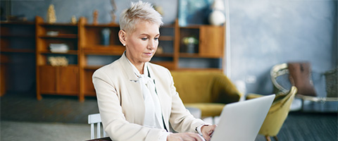 A person with short light hair wearing a cream blazer sits in a chair in a warmly decorated space, typing on a laptop with a focused expression. A bookshelf, yellow armchair, and plants fill the background.