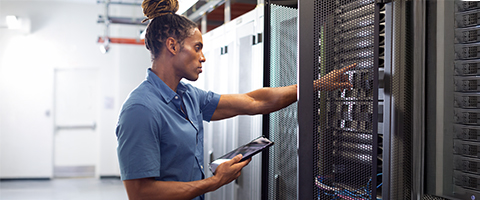 A person with hair tied back, wearing a blue polo shirt, holds a tablet while reaching toward a row of tall server racks in a data center.