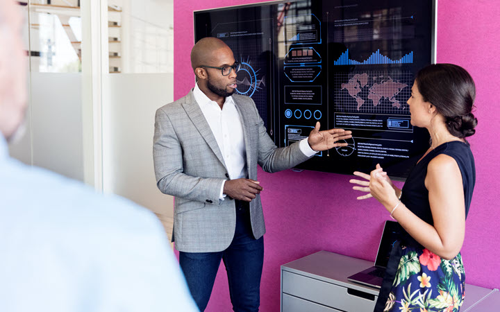 A professional setting featuring a man in a gray blazer presenting data on a large digital screen. The screen displays graphs, charts, and a world map, suggesting analytics or global trends. The background includes a vibrant pink wall and office furniture, creating a modern workspace atmosphere.