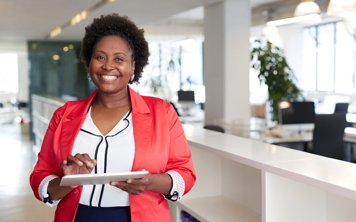 A woman stands in a modern office environment holding and interacting with a tablet device. The setting features bright lighting, open workspaces, and indoor plants, creating a professional and welcoming atmosphere. She is dressed in a red blazer and white blouse, adding a touch of vibrant style to the scene.
