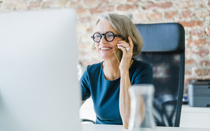 A smiling woman, in business attire, talking on a phone while sitting at a desk.