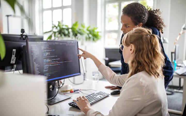 Two women in an office working on a computer. One woman is seated and pointing at the computer screen. The other is standing and looking at the screen.