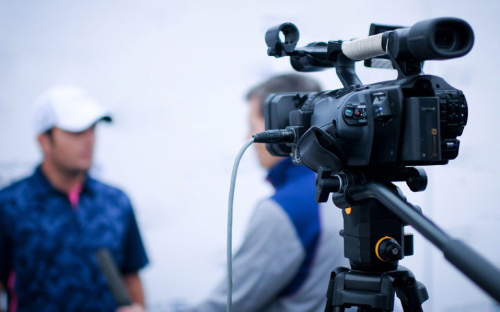 A video camera is visible in the foreground while two men talk in the background.