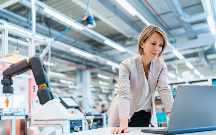 A woman in business attire looks at a laptop screen. The laptop is laying on a desk in a modern factory.