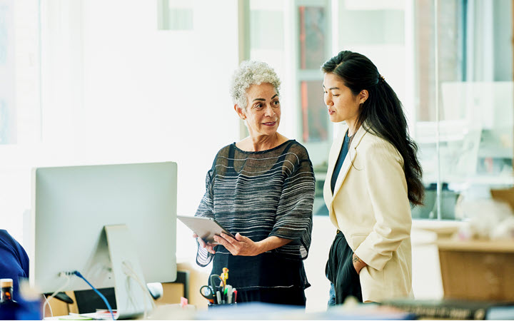 Two women stand at a desk in an office. One woman is holding a tablet and the other woman is looking at the screen.