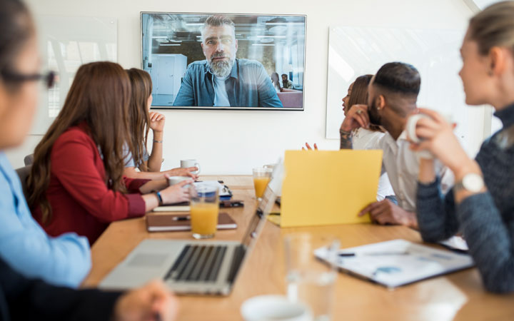 Workers in an office sit around a conference table while looking at a man talking on a television monitor