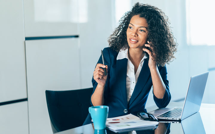 A woman in an office holding a pen and talking on a mobile phone. There is a laptop and paperwork on her desk.