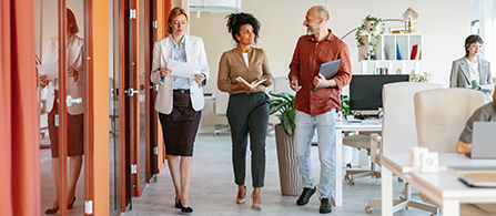 Three business colleagues walking down a well light open-air office space carrying laptops and notebook having a lively discussion.