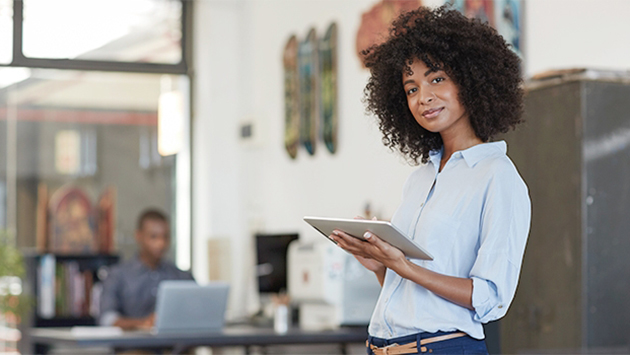 A person with curly hair, wearing a light blue button-up shirt, stands in an office holding a tablet in both hands and looks toward the camera with a slight smile. In the softly blurred background, another person sits working at a desk with a laptop, and framed artwork hangs on the wall.