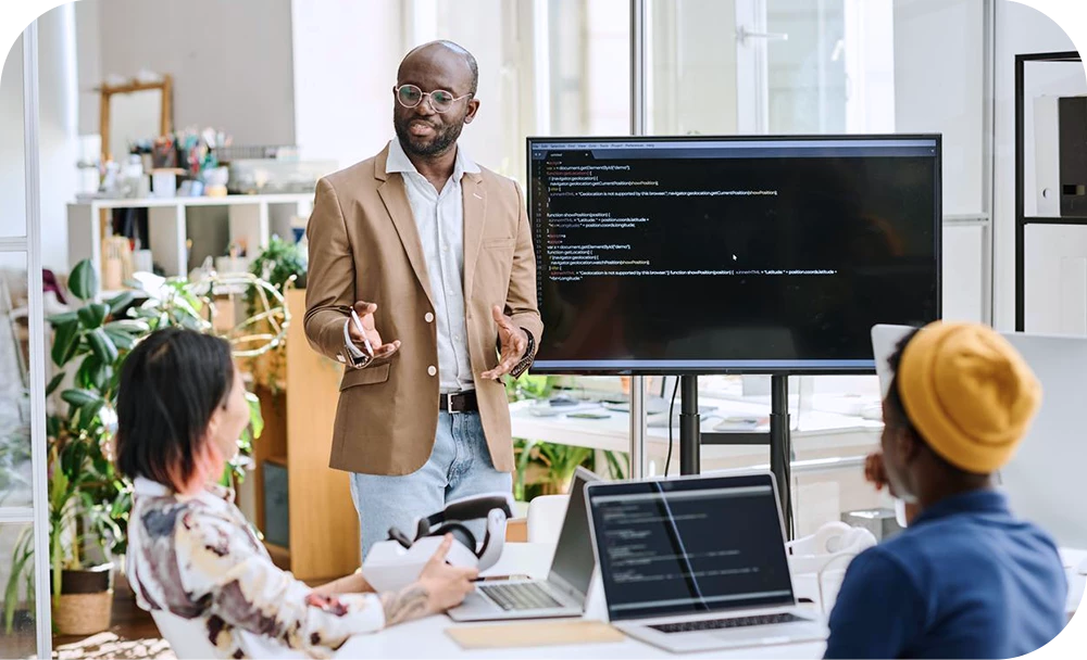 A person in a tan blazer stands and gestures toward a large monitor displaying lines of code while presenting to two colleagues seated in the foreground. The setting is a bright, plant-filled office with shelves and greenery.