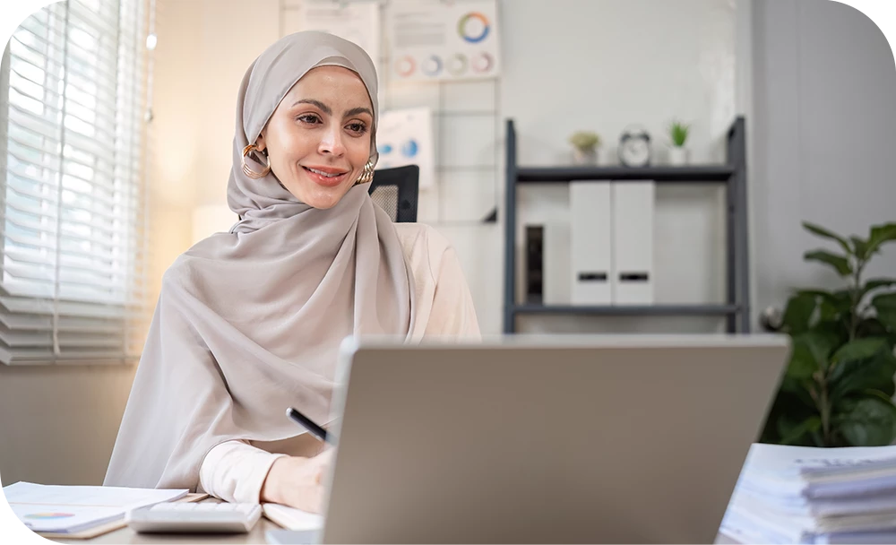 A person wearing a headscarf and a cream blazer sits at a desk in a bright office, smiling toward the camera with a laptop open in front of them. A stack of papers and binders on a shelf are visible in the softly blurred background.