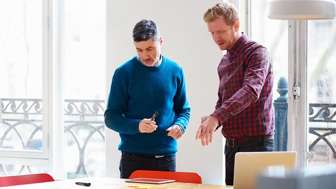Two people in business casual dress, one in a blue sweater and jeans and the other in a red and blue gingham button-up and jeans, stand in conversation while looking down at a tablet on a large wooden table. 