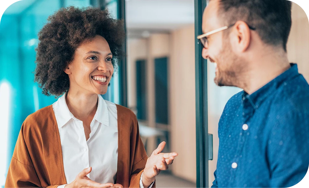 Two people stand close together in a bright office hallway smiling and talking. One in a rust-colored cardigan gestures while speaking, and the other in a blue shirt and glasses listens and smiles.