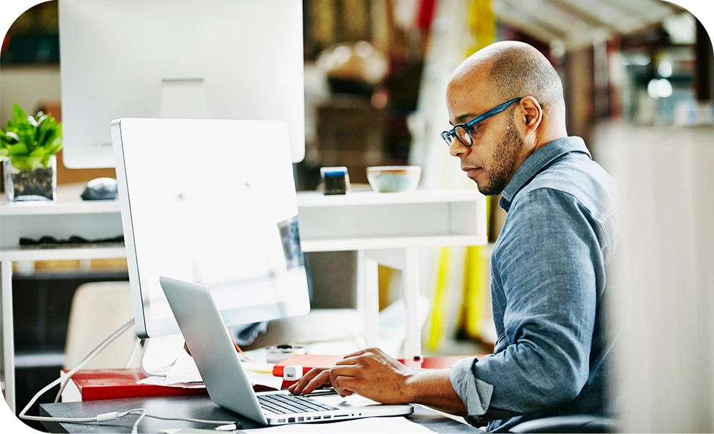 A person in a gray sweater and glasses sits in profile at a desk in a bright studio-like office, typing on a laptop with a focused expression. Shelves and plants are visible in the softly blurred background.