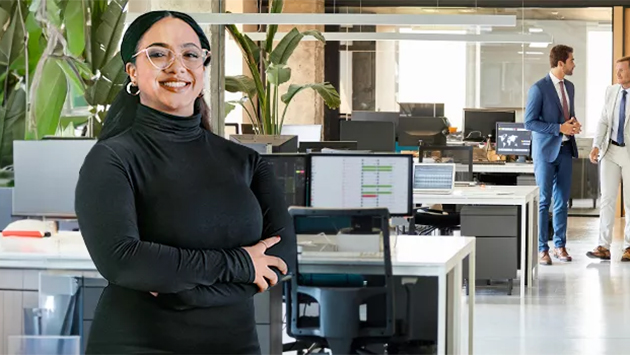 A person with glasses and a head covering, wearing a black turtleneck, stands with arms crossed and a warm smile in a large modern open-plan office. Behind them, two other people stand talking near desks with monitors and potted plants.