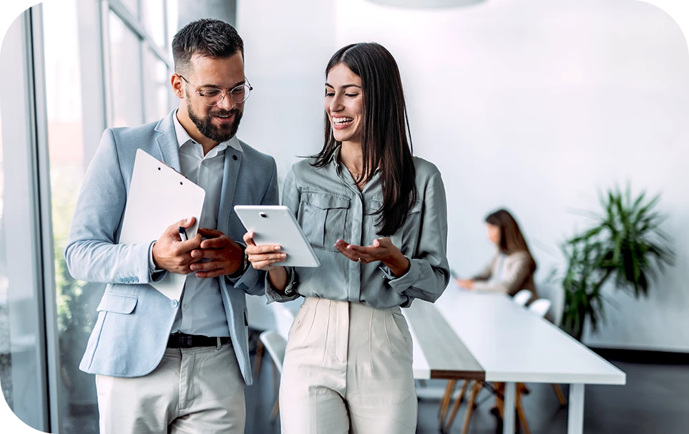 Two people walk side by side in a bright office, smiling and looking at a tablet one of them holds. One wears a light gray blazer and glasses; the other wears a gray shirt and jeans. A colleague works at a desk in the blurred background.