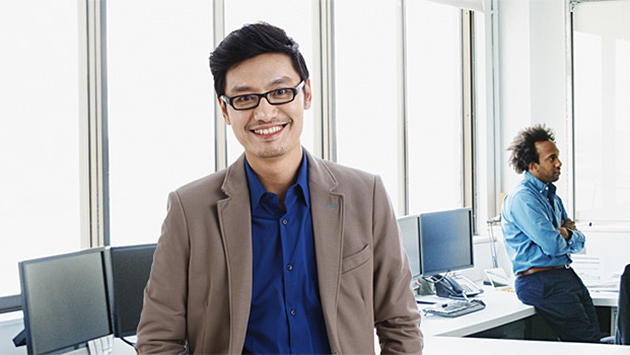 A person with glasses in a tan blazer over a blue shirt stands in the foreground of a bright open office, smiling toward the camera. A colleague leans against a desk with arms crossed near large windows in the background.