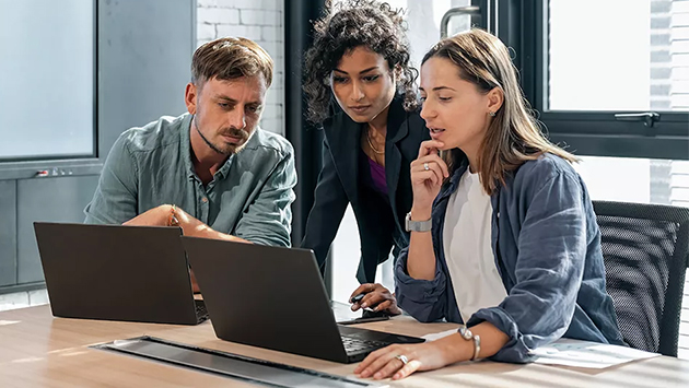 Three people gather around two laptops at a table in a modern office, looking intently at the screens. One person is seated on the left in a gray shirt, another stands in the center in a dark blazer leaning in, and a third sits on the right in a denim jacket with a hand resting near their chin. An exposed brick wall and windows are visible in the background.