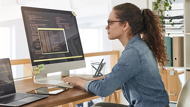 A person with curly hair tied back, wearing glasses and a denim shirt, sits in profile at a wooden desk typing on a keyboard while looking at a large desktop monitor that displays lines of code on a dark screen. A laptop, smartphone, and sticky notes are on the desk, in a bright office with plants and shelving in the background.