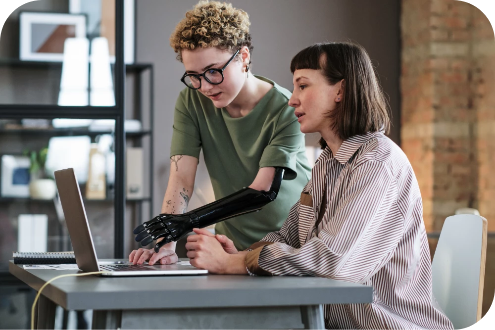 Two people look at a laptop together at a desk. One stands and leans in wearing a green T-shirt and glasses, pointing toward the screen; the other sits in a light blouse, focused on the laptop. The setting is a loft-style office with exposed brick.