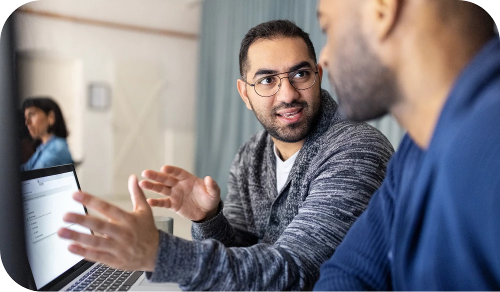 A person wearing glasses and a striped sweater leans forward and gestures with both hands while talking to a colleague across a table, with an open laptop between them in a bright office. Another person sits at a desk in the blurred background.
