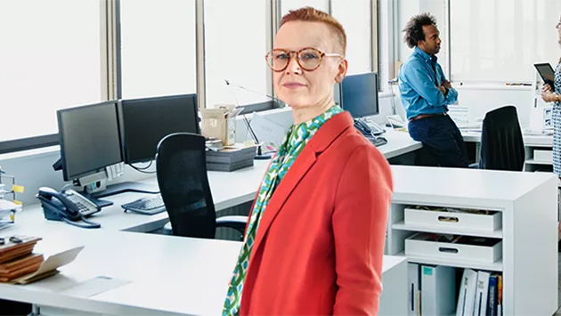 A person with short reddish hair, glasses, an orange-red blazer, and a green patterned scarf stands smiling in an open office lined with desks and monitors. Two colleagues stand talking near the windows in the background.