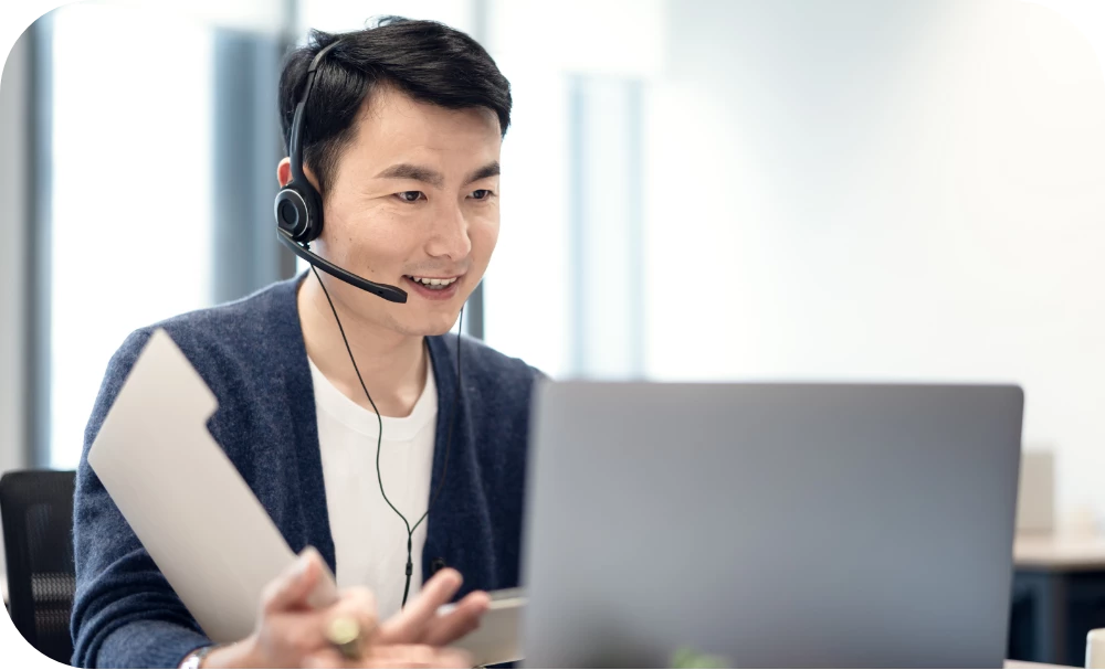 A person wearing a headset and a dark cardigan over a white shirt sits at a desk, smiling while looking at an open laptop and holding papers. A bright office with large windows is softly blurred in the background.