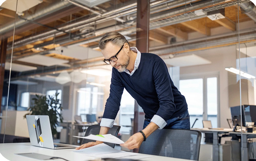 A person with gray hair and glasses, wearing a dark sweater, stands leaning over a desk reviewing papers beside an open laptop in an open-plan office with exposed ceiling ductwork and large windows.