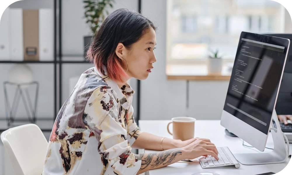 A person with dark hair ending in pink tips, wearing a floral-patterned top, sits at a desk in a bright home office typing on a desktop computer. A coffee mug sits beside the keyboard, with soft greenery and shelving blurred in the background.